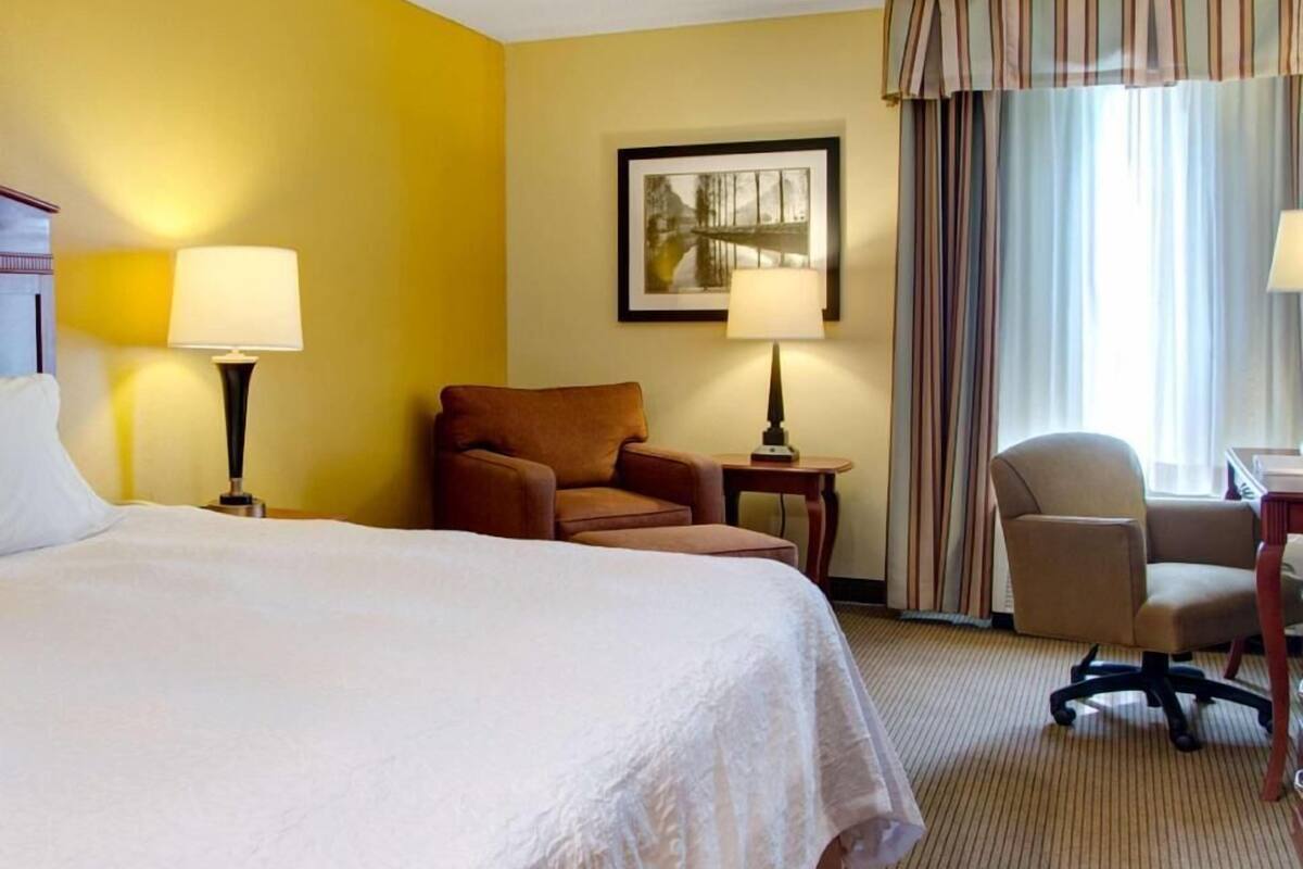A hotel room with a bed, two chairs, a desk, a lamp, a framed picture on the wall, and a window with curtains.