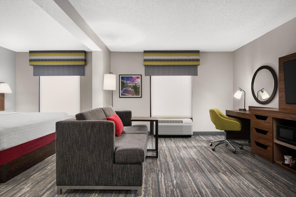 A hotel room with a bed, a gray sofa with red cushions, a desk with a chair, a large mirror, and an air conditioning unit.
