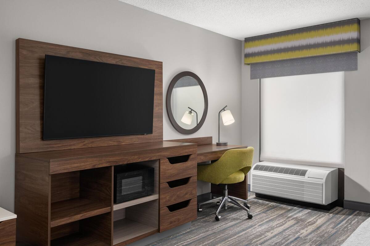 A modern hotel room with a large flat-screen TV mounted on a wooden cabinet, a round mirror above it, a desk with a chair, and a large window with a view.