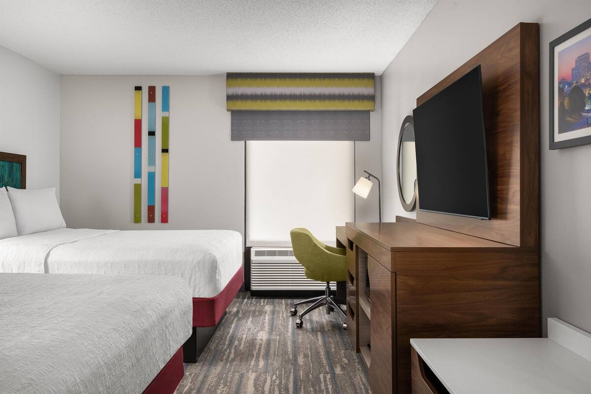 A hotel room with two beds, a desk with a chair, a flat-screen TV, a window with a view, and a wall-mounted air conditioner.