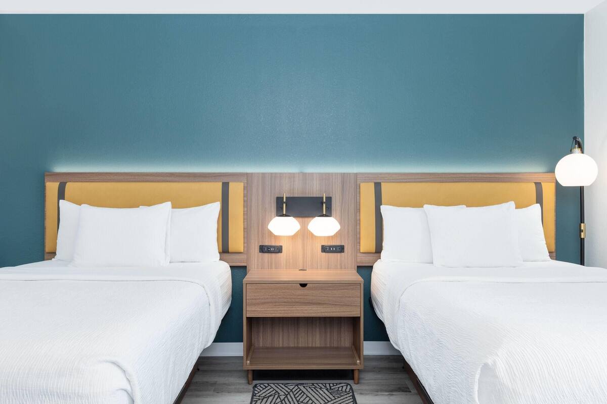 A hotel room with two beds, a wooden headboard, a nightstand with two lamps, and a patterned rug.