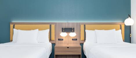 A hotel room with two beds, a wooden headboard, a nightstand with two lamps, and a patterned rug.