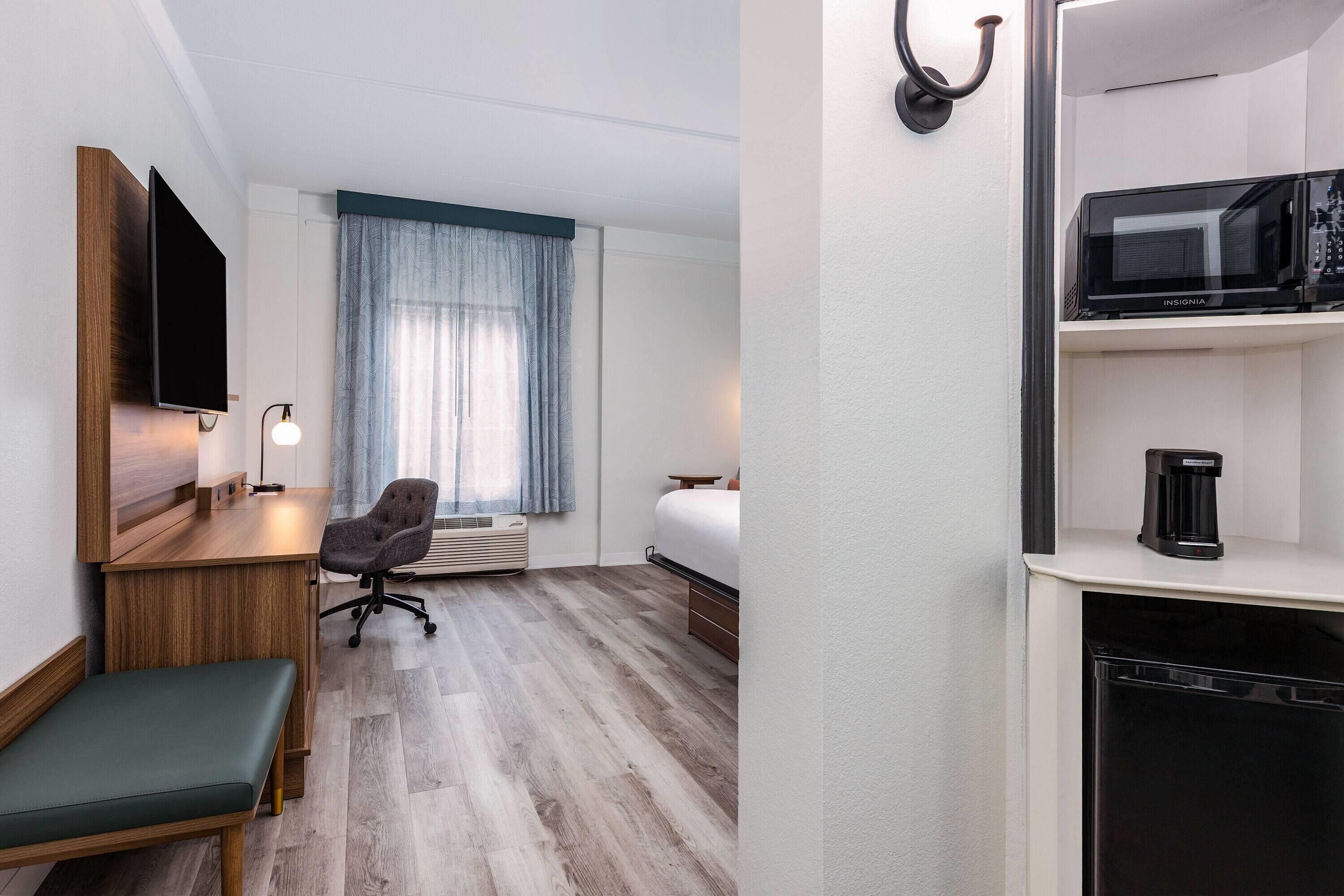 Habitación de hotel con escritorio de madera, un banco con cojín verde, una silla de oficina gris, una cama, una ventana con cortinas azules, un televisor de pantalla plana montado en la pared, un microondas y una cafetera.