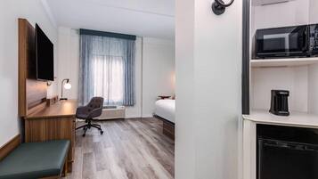 Habitación de hotel con escritorio de madera, un banco con cojín verde, una silla de oficina gris, una cama, una ventana con cortinas azules, un televisor de pantalla plana montado en la pared, un microondas y una cafetera.