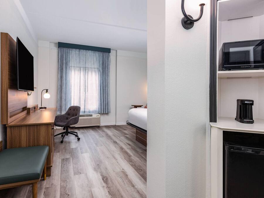 Habitación de hotel con escritorio de madera, un banco con cojín verde, una silla de oficina gris, una cama, una ventana con cortinas azules, un televisor de pantalla plana montado en la pared, un microondas y una cafetera.