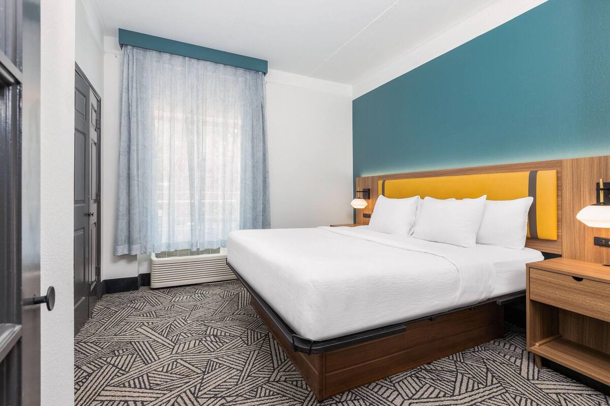 A hotel room with a bed, a nightstand, a window with curtains, and a patterned floor.