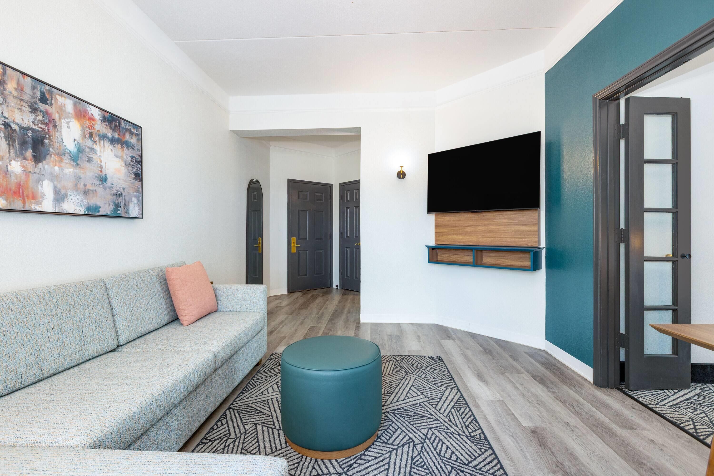 A modern living room with a grey sofa, teal ottoman, abstract painting, and a mounted TV.