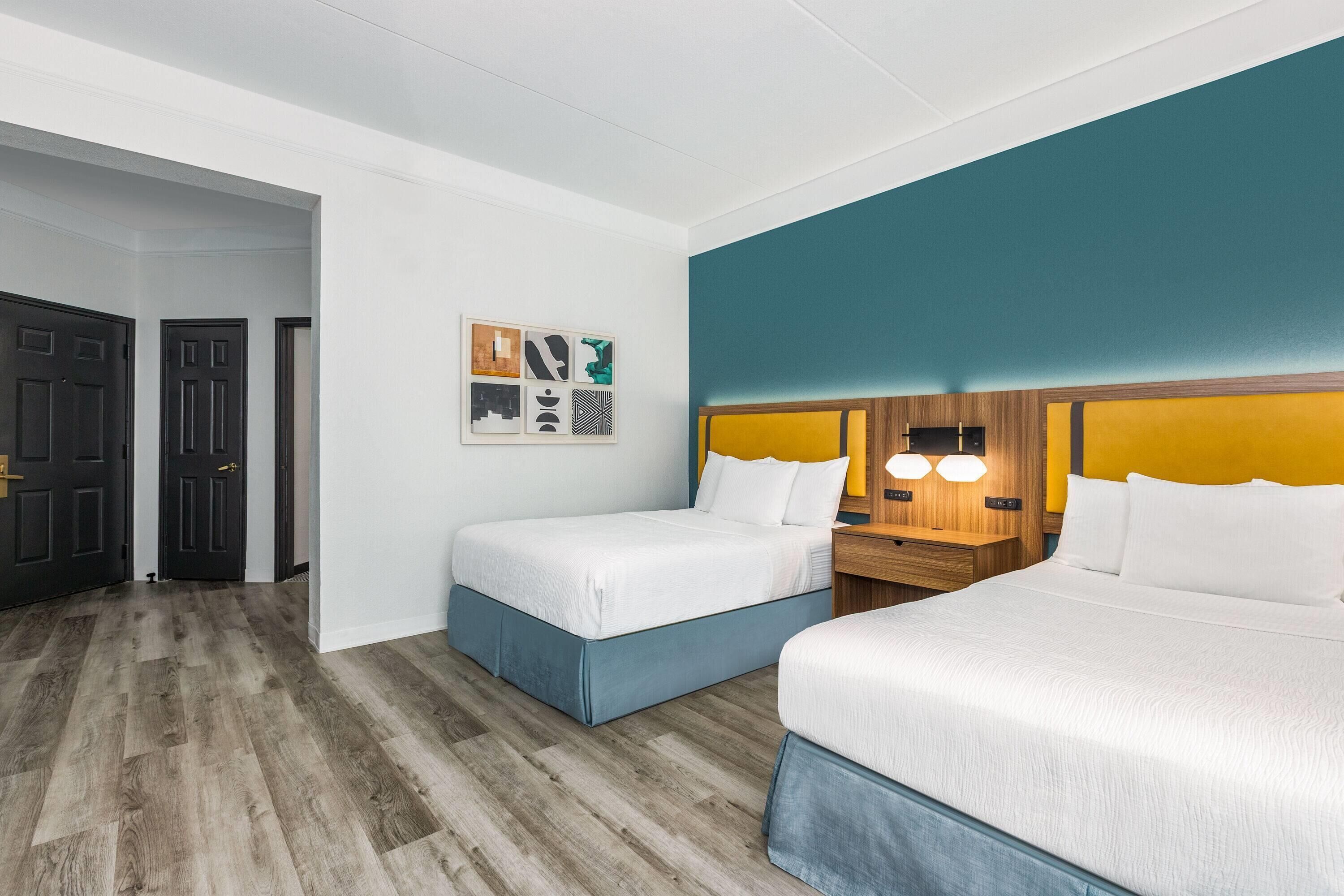 A hotel room with two beds, a wooden floor, a white wall with a teal accent, and a small table with a lamp.