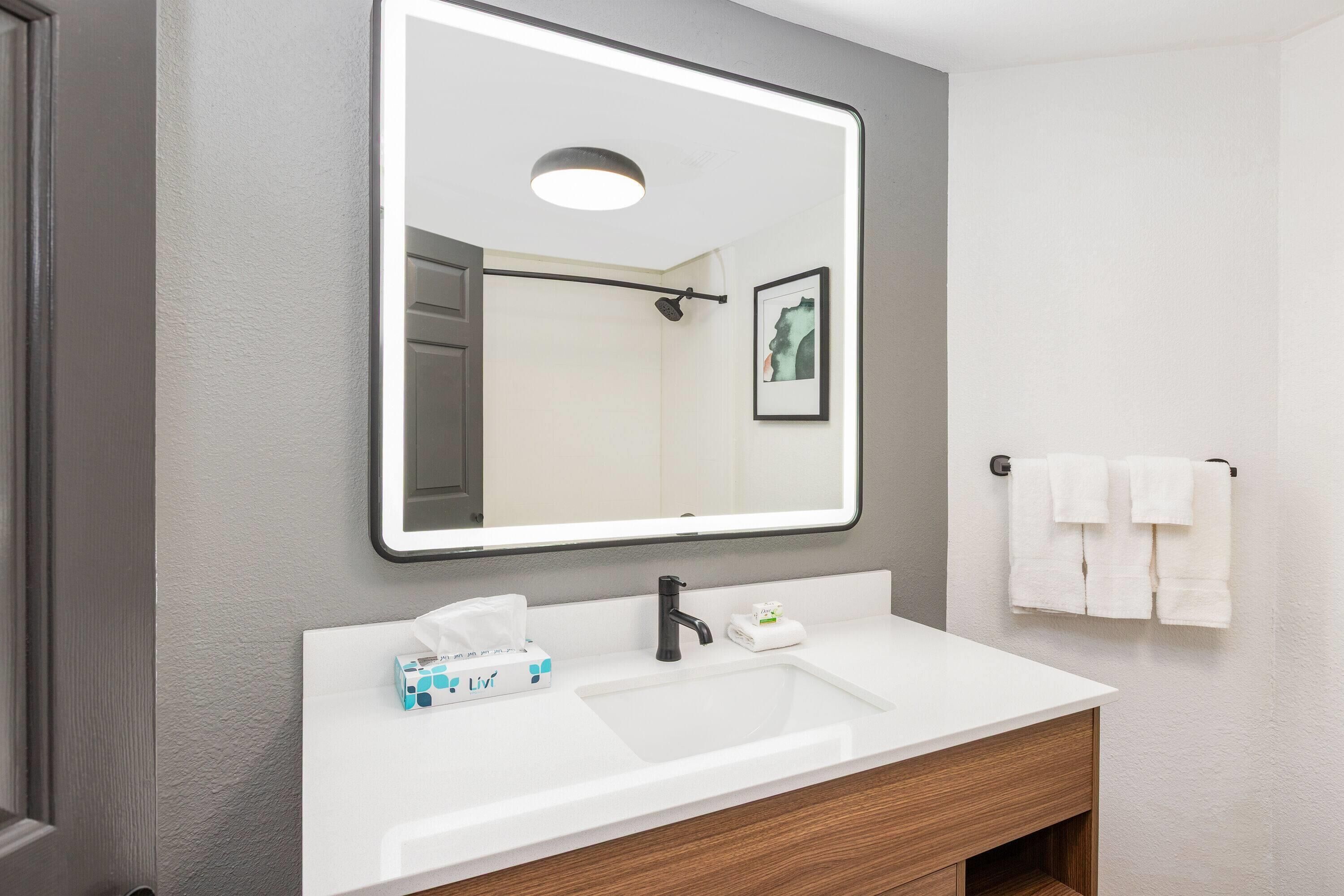 A modern bathroom with a white sink, black faucet, and a large mirror. There is a framed artwork on the wall and a towel rack with white towels.