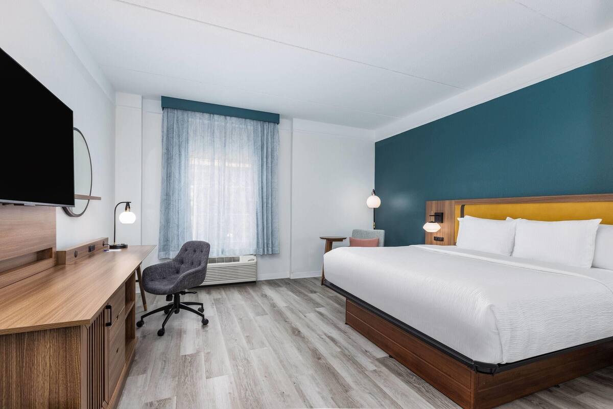 A hotel room with a wooden desk, a comfortable chair, a large bed with white linens, a television on the wall, and a window with sheer curtains.