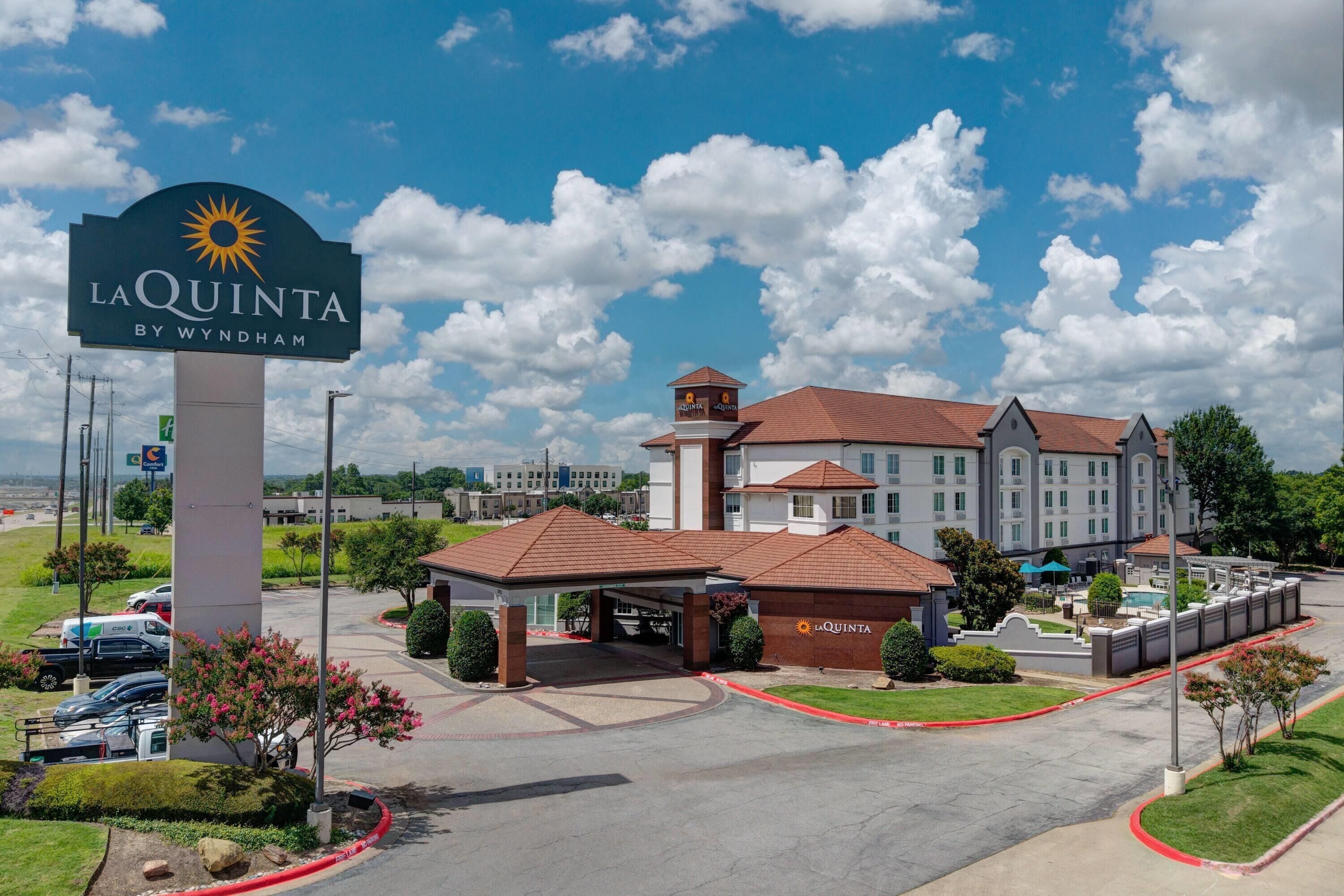 A hotel with a large sign reading "La Quinta by Wyndham," featuring a sun emblem. It has a covered entrance, a swimming pool, and a well-maintained lawn.