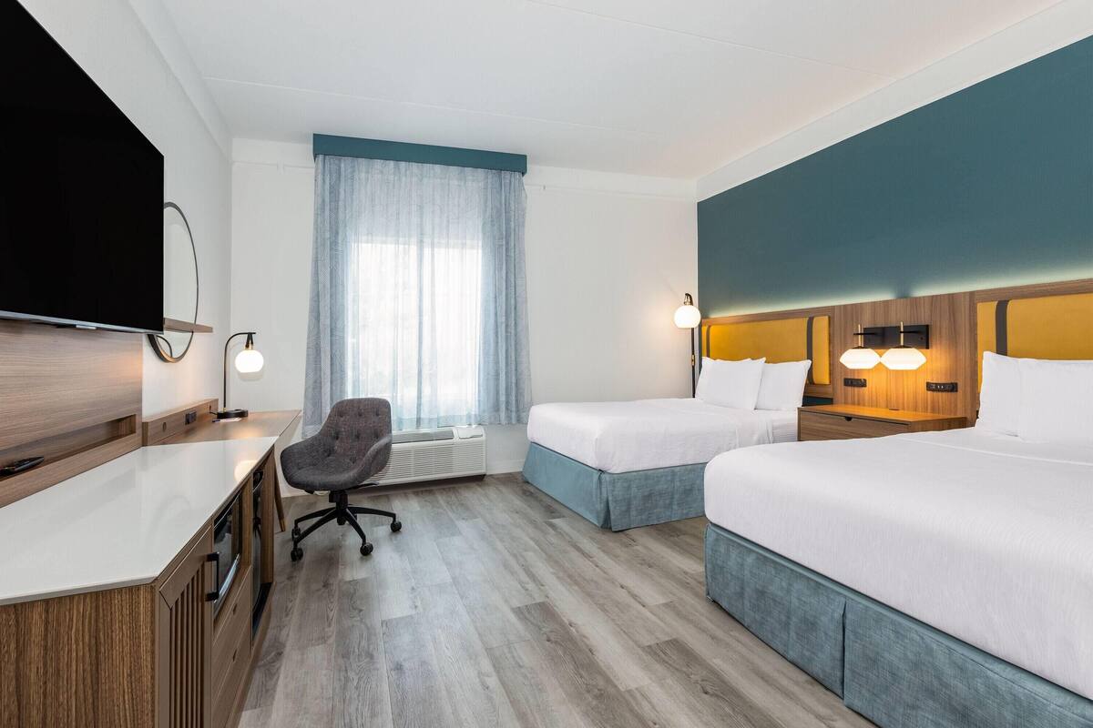 A hotel room with a desk, two beds, a TV, a chair, and a window with curtains.