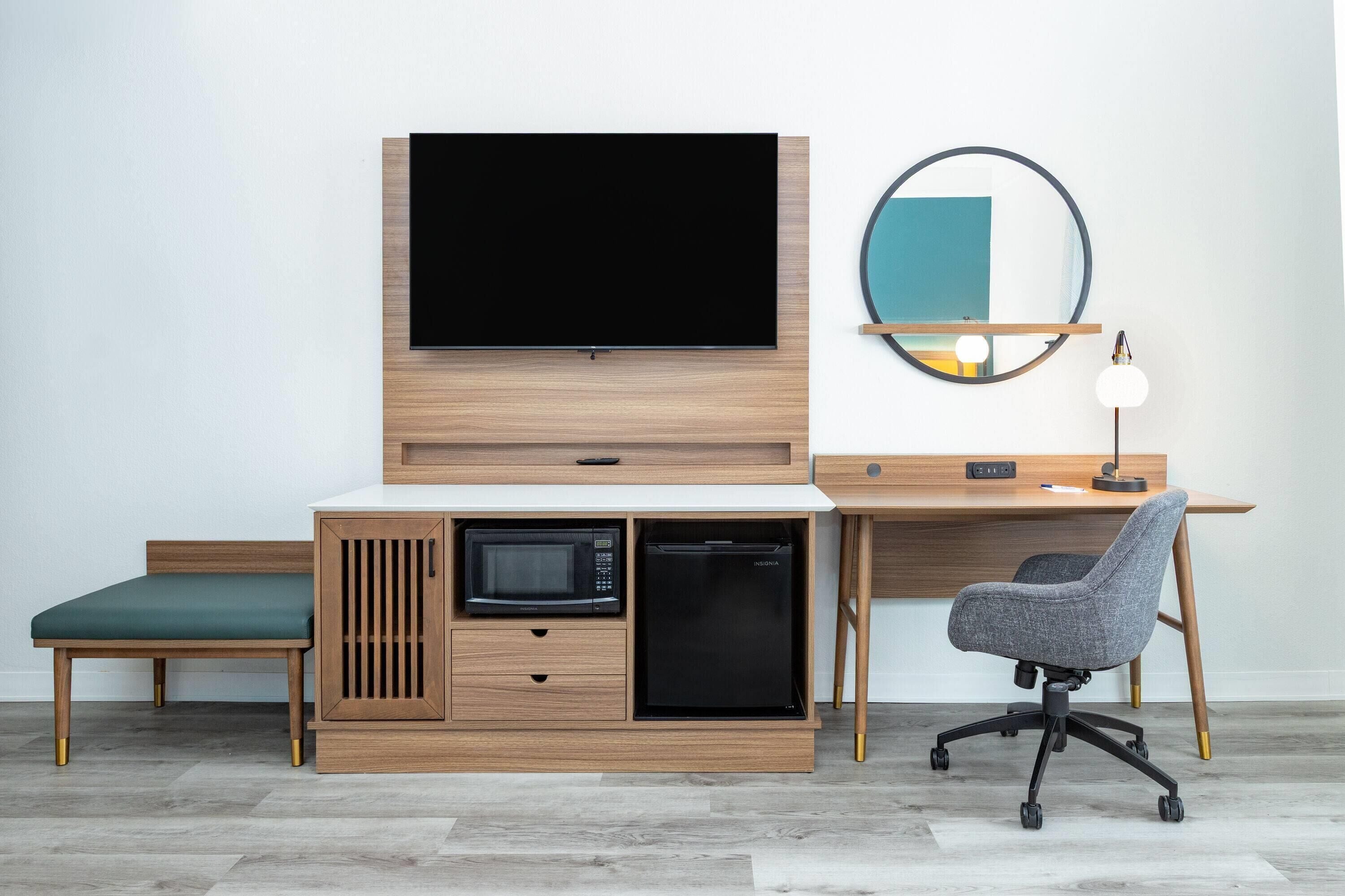 Una habitación moderna con un escritorio de madera, televisor, microondas, refrigerador, una silla y un espejo redondo en la pared.