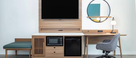 A modern room with a wooden desk, a television, a microwave, a refrigerator, a chair, and a round mirror on the wall.