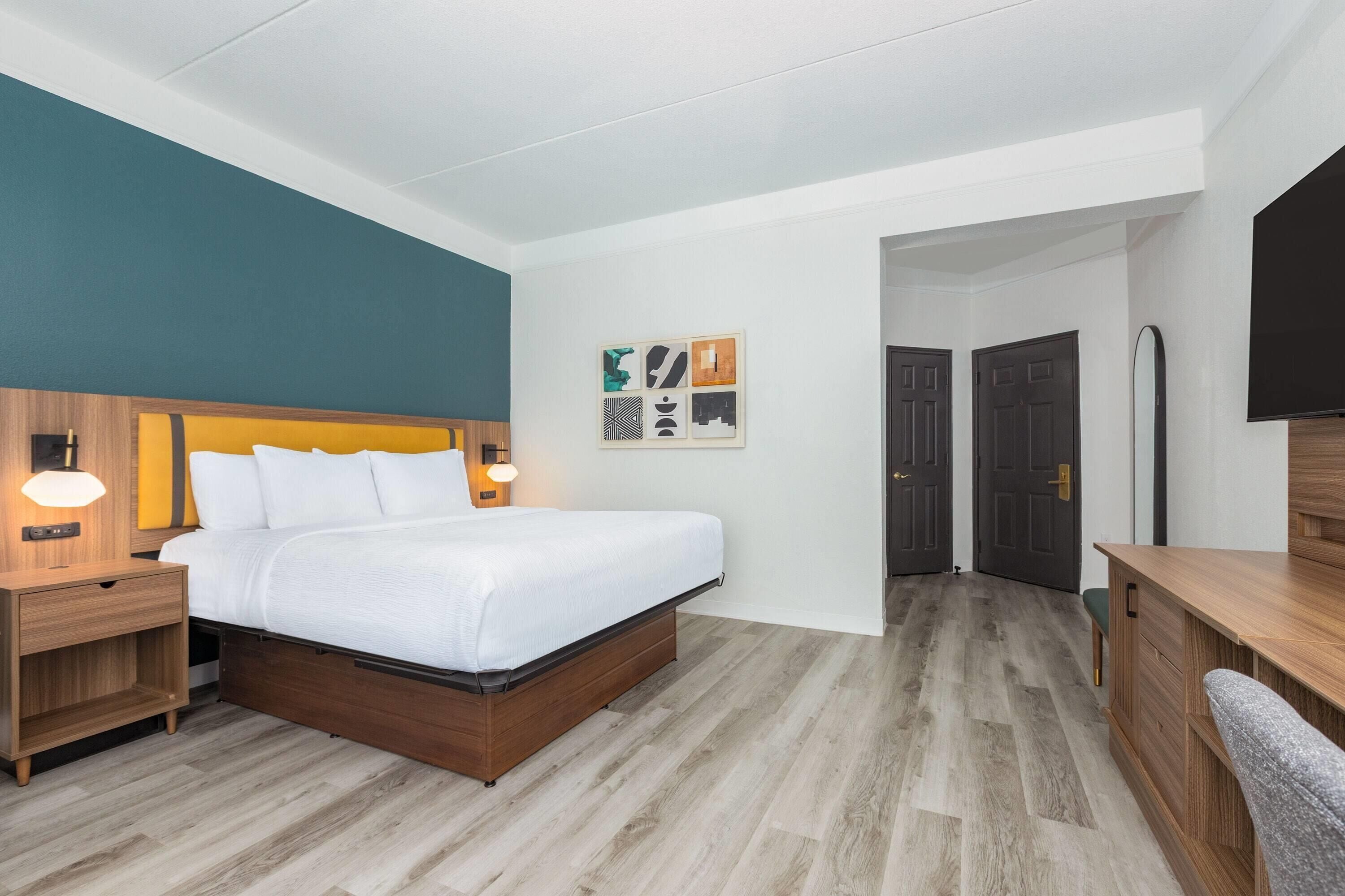 Een hotelkamer met een bed, nachtkastjes, een bureau en een stoel. Er zijn deuren die leiden naar andere kamers en een gang.