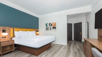 A hotel room with a bed, bedside tables, a desk, and a chair. There are doors leading to other rooms and a hallway.
