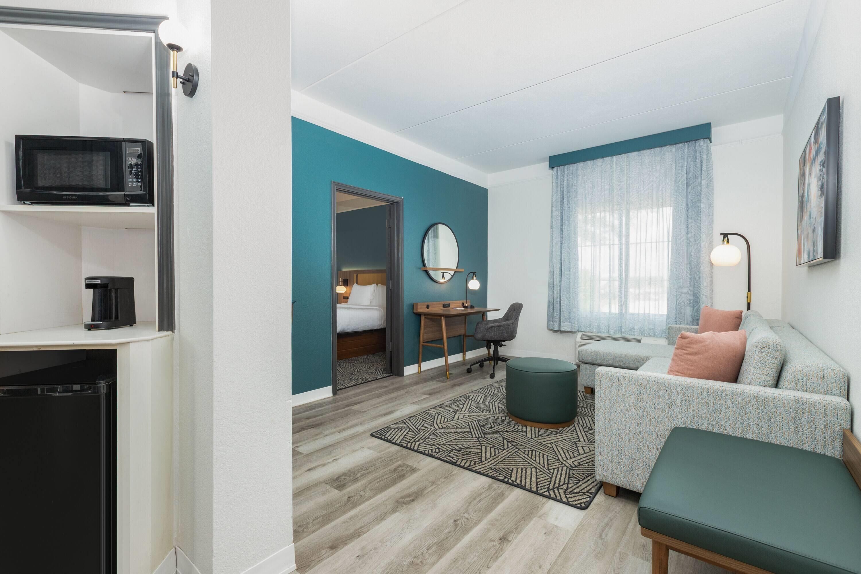 Une chambre d’hôtel compacte comprenant un coin cuisine, un lit, un bureau, un canapé et une grande fenêtre avec des rideaux.