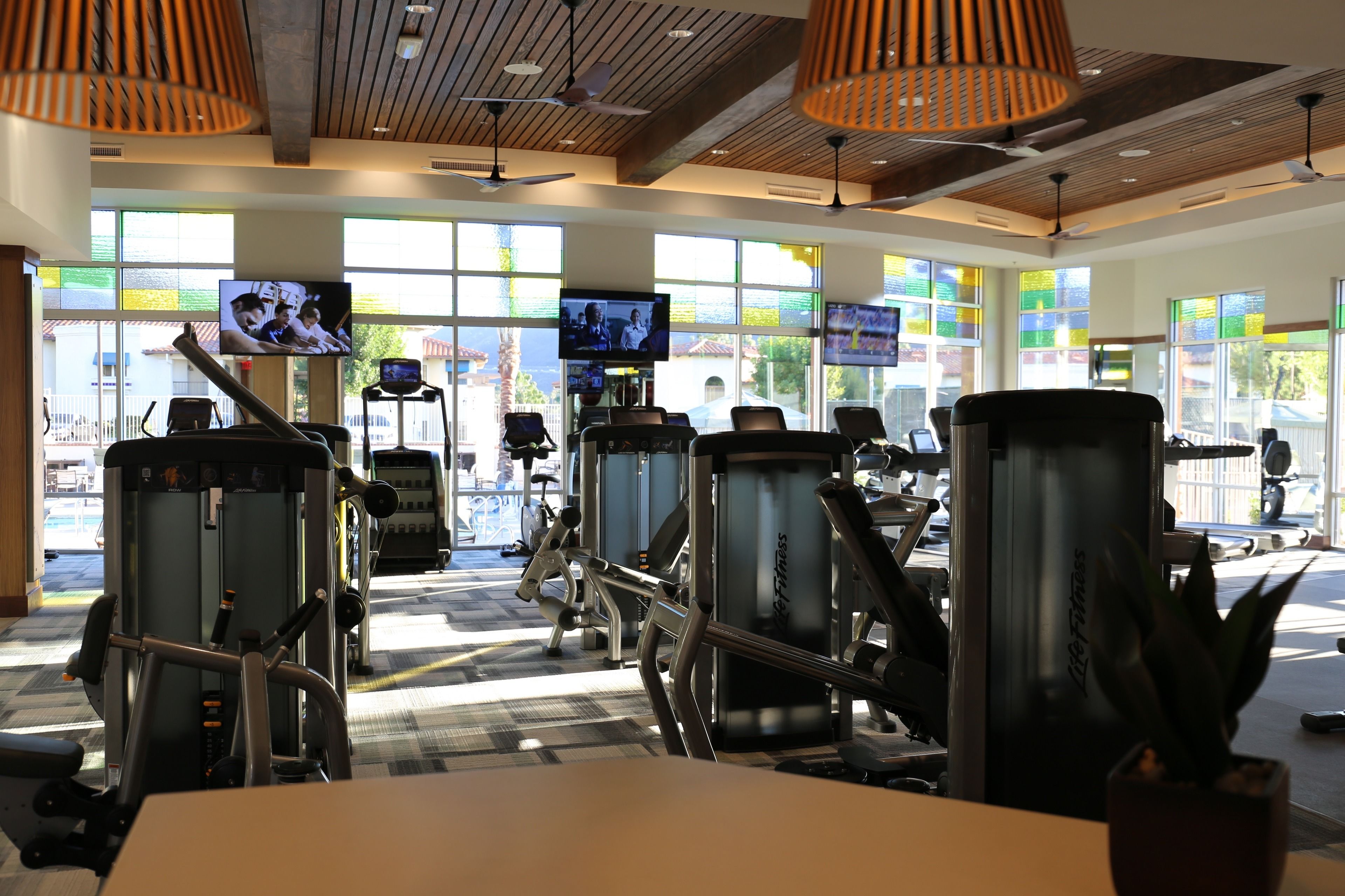 A gym with various exercise machines, large windows, and multiple televisions displaying content.