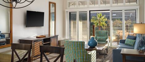 A well-lit living room with a TV, seating area, and large windows with blinds.