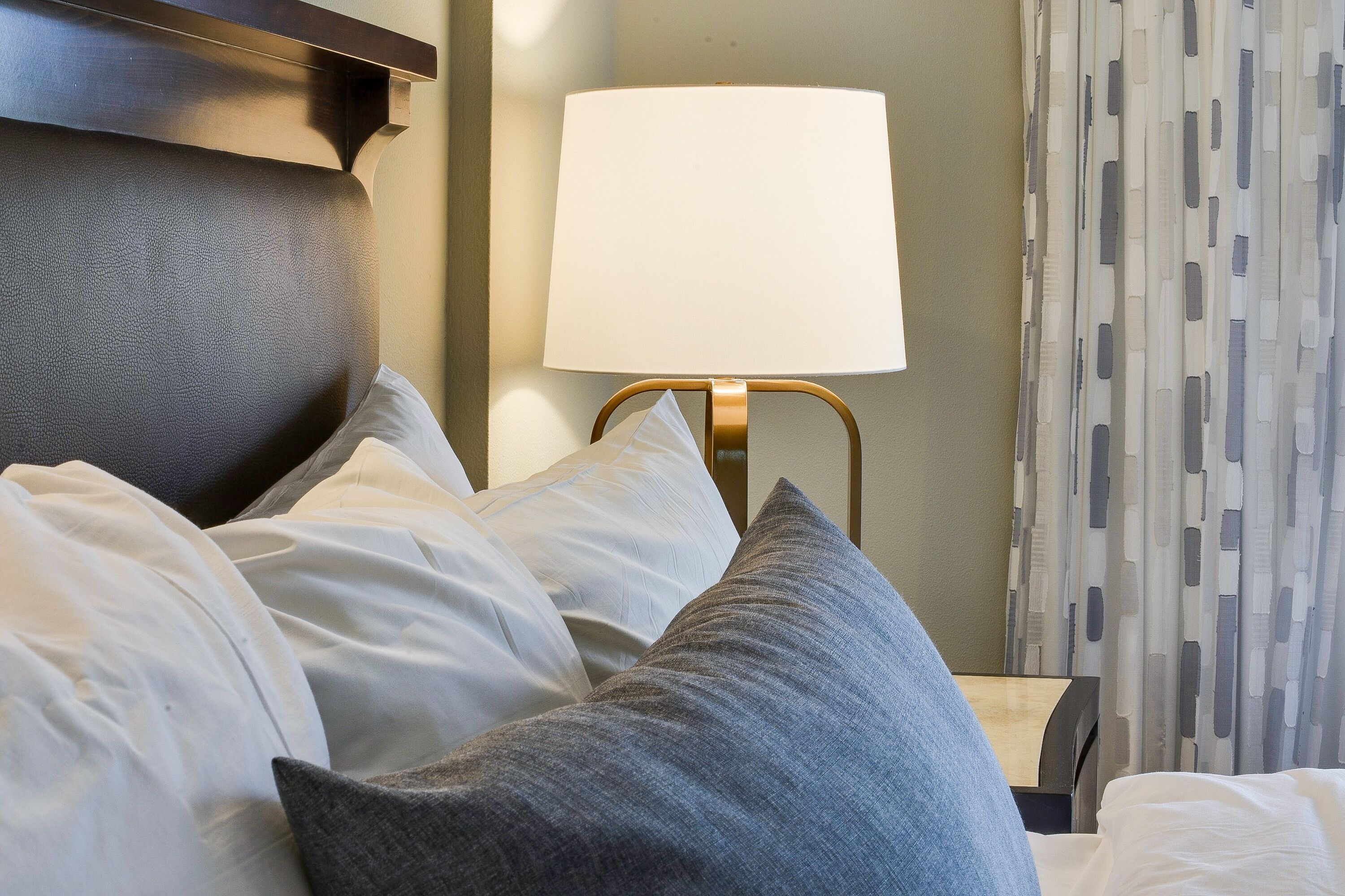 A neatly made bed with white and gray pillows, a gold-framed lamp, and a patterned curtain in a hotel room.