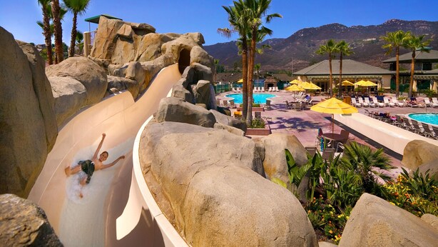 A water slide with large rocks, a pool area with lounge chairs and umbrellas, and mountains in the background. - Hyatt Vacation Club at The Welk, San Diego Area (Escondido)