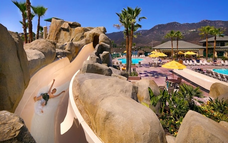 A water slide with large rocks, a pool area with lounge chairs and umbrellas, and mountains in the background.