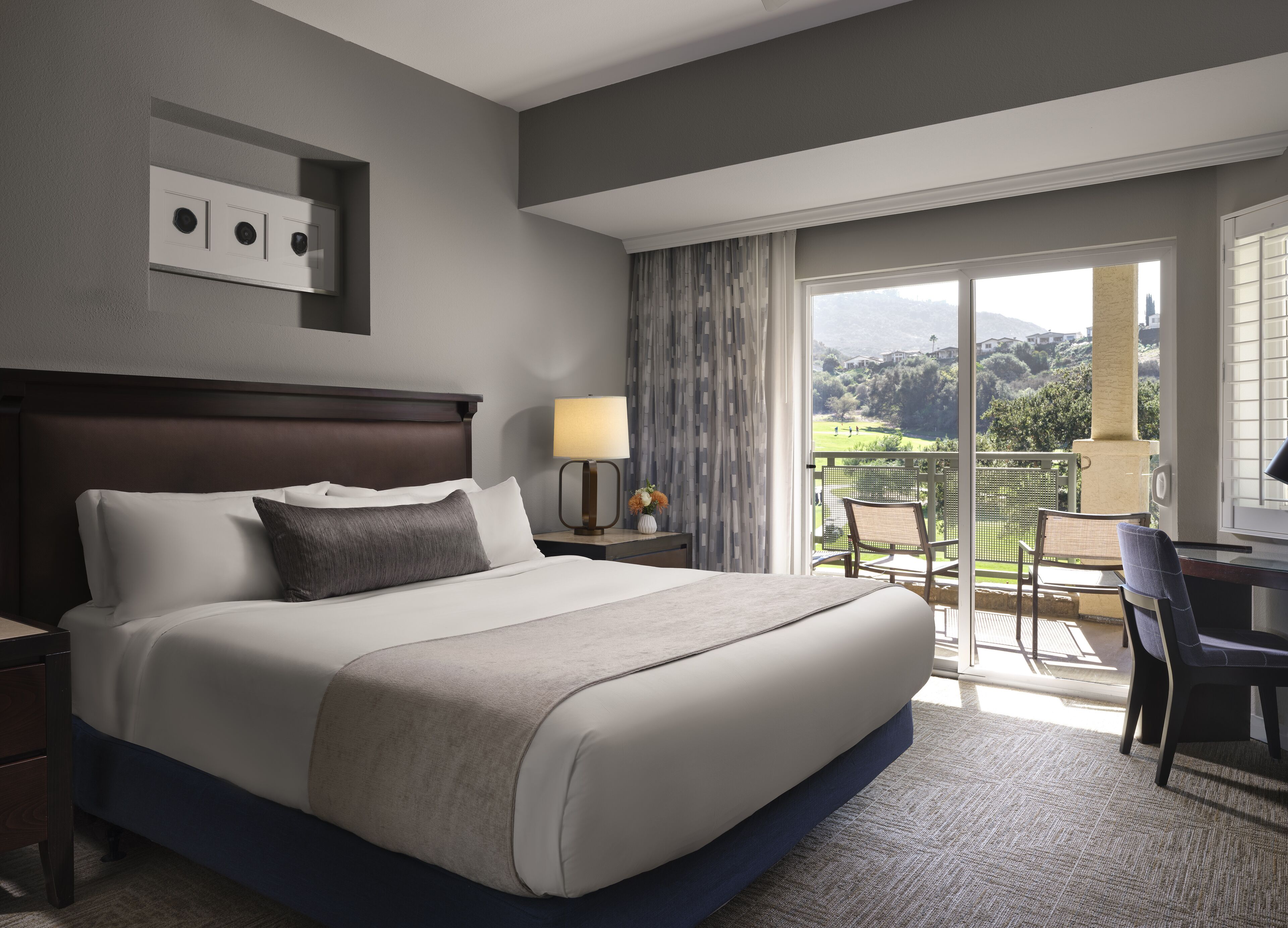 Une chambre d’hôtel avec un grand lit, une table de chevet, une lampe, une chaise et une vue sur un balcon donnant sur un paysage pittoresque.