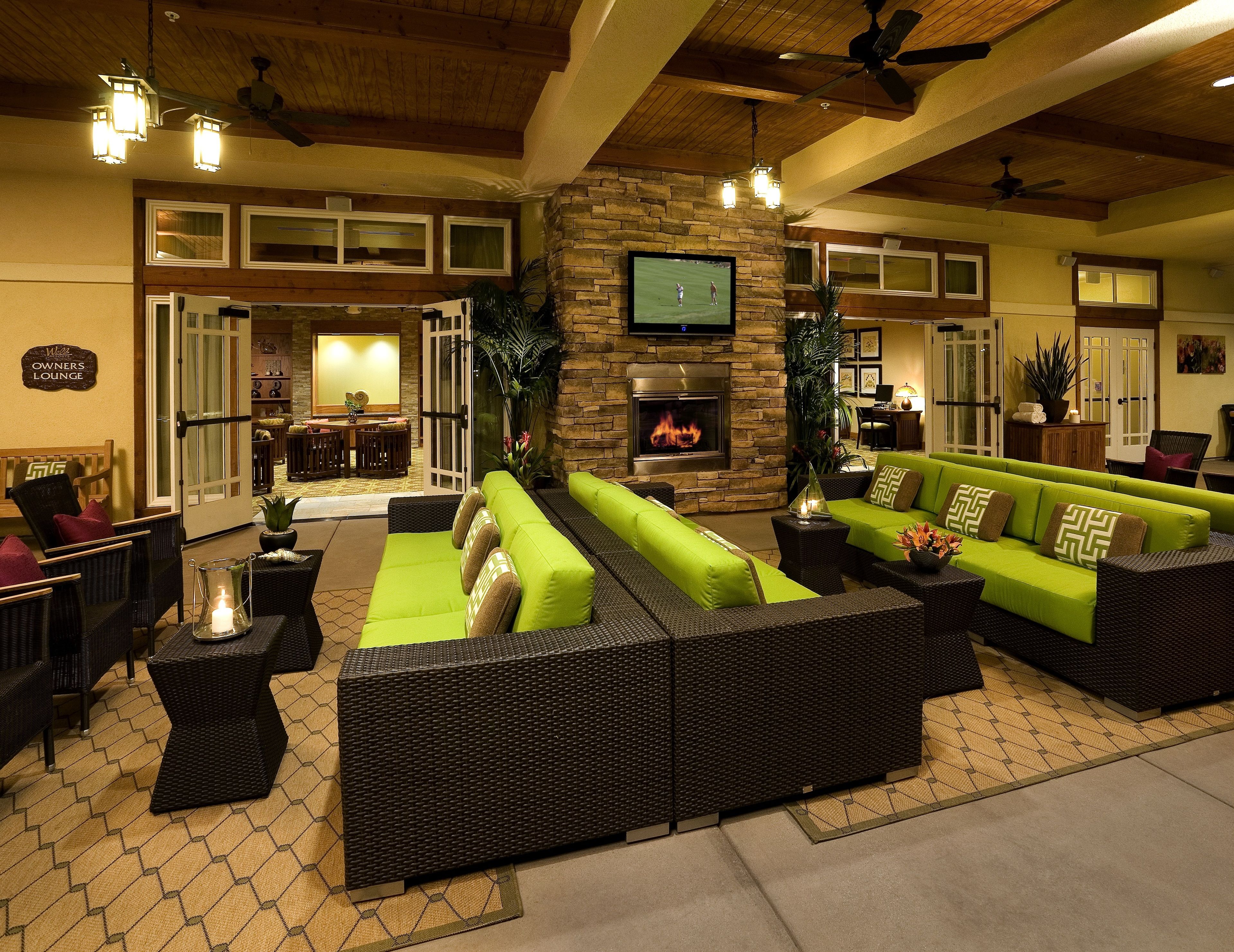 A spacious lounge area with green sofas, wicker furniture, a stone fireplace, and a television mounted on the wall.