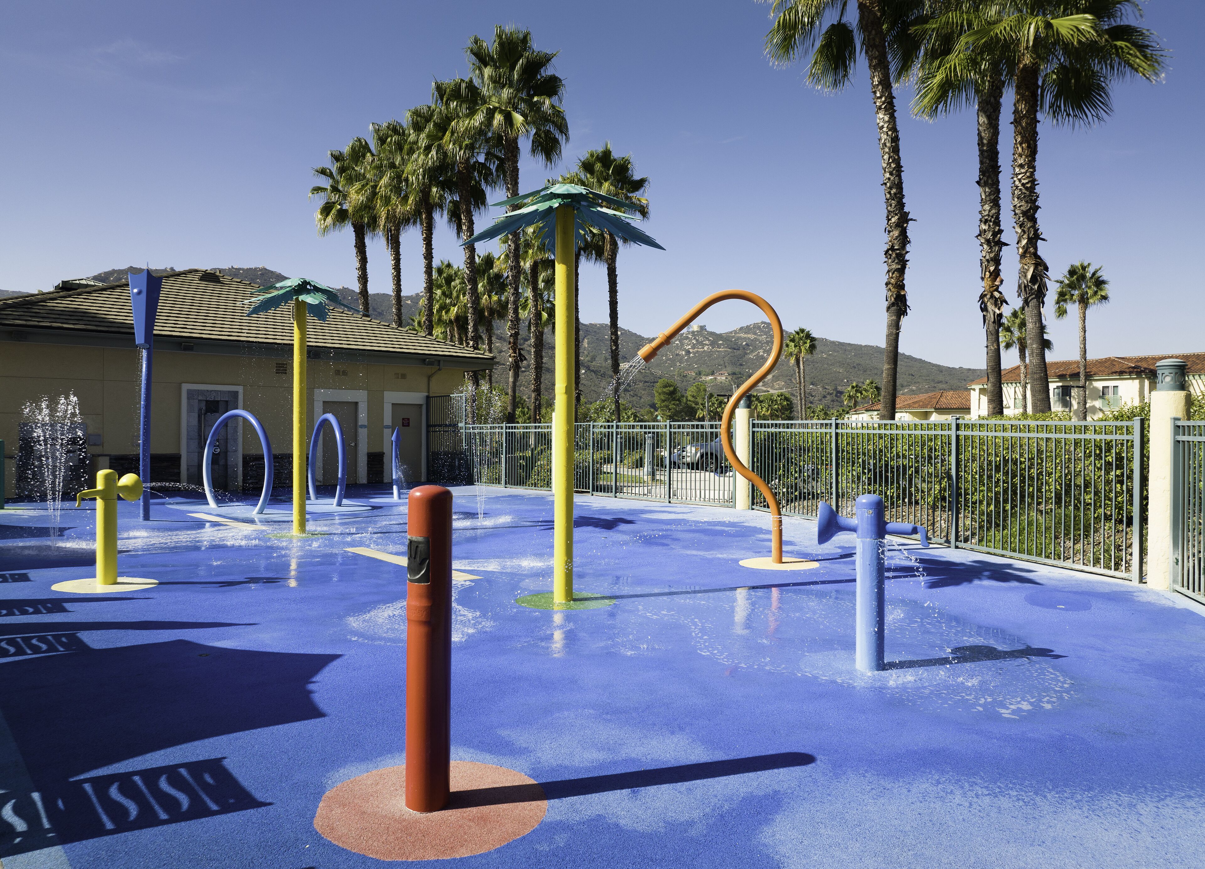 An outdoor water play area with a blue rubber surface, water jets, and a yellow pole.