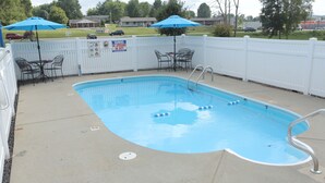 A rectangular pool with a metal ladder, surrounded by a white fence. There are two blue umbrellas and a table with chairs.