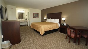 A hotel room with a bed, bedside table, desk, chair, and a small trash bin.