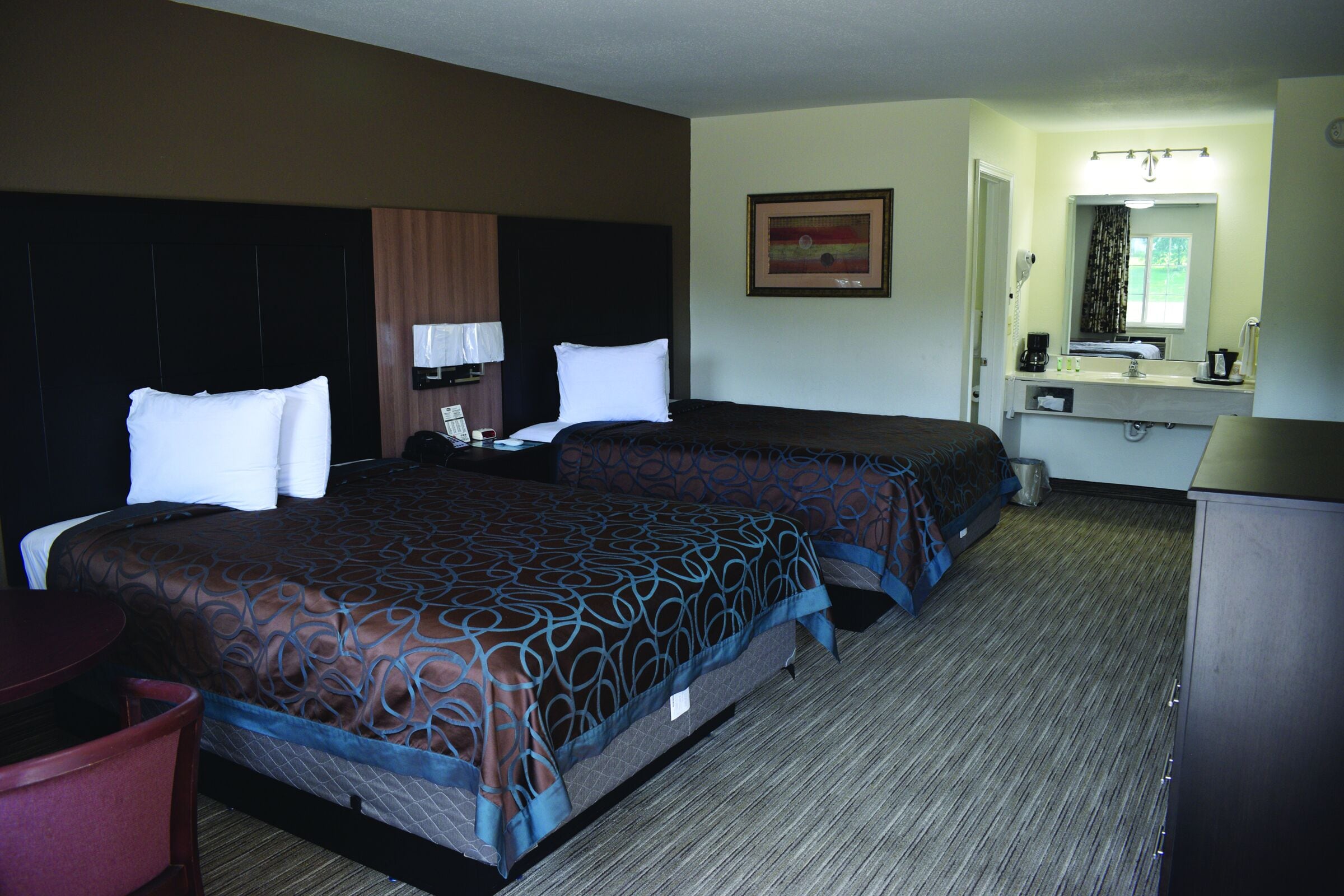 A hotel room with two beds, a desk, a chair, a mirror, a sink, and a window.