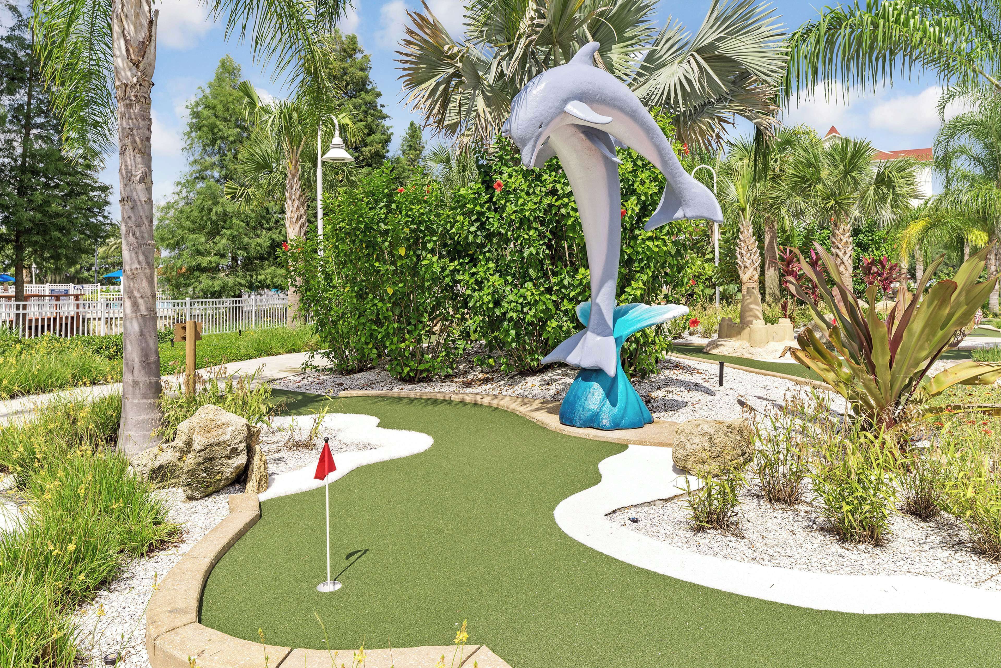 A miniature golf course with a dolphin sculpture, surrounded by tropical plants and palm trees.