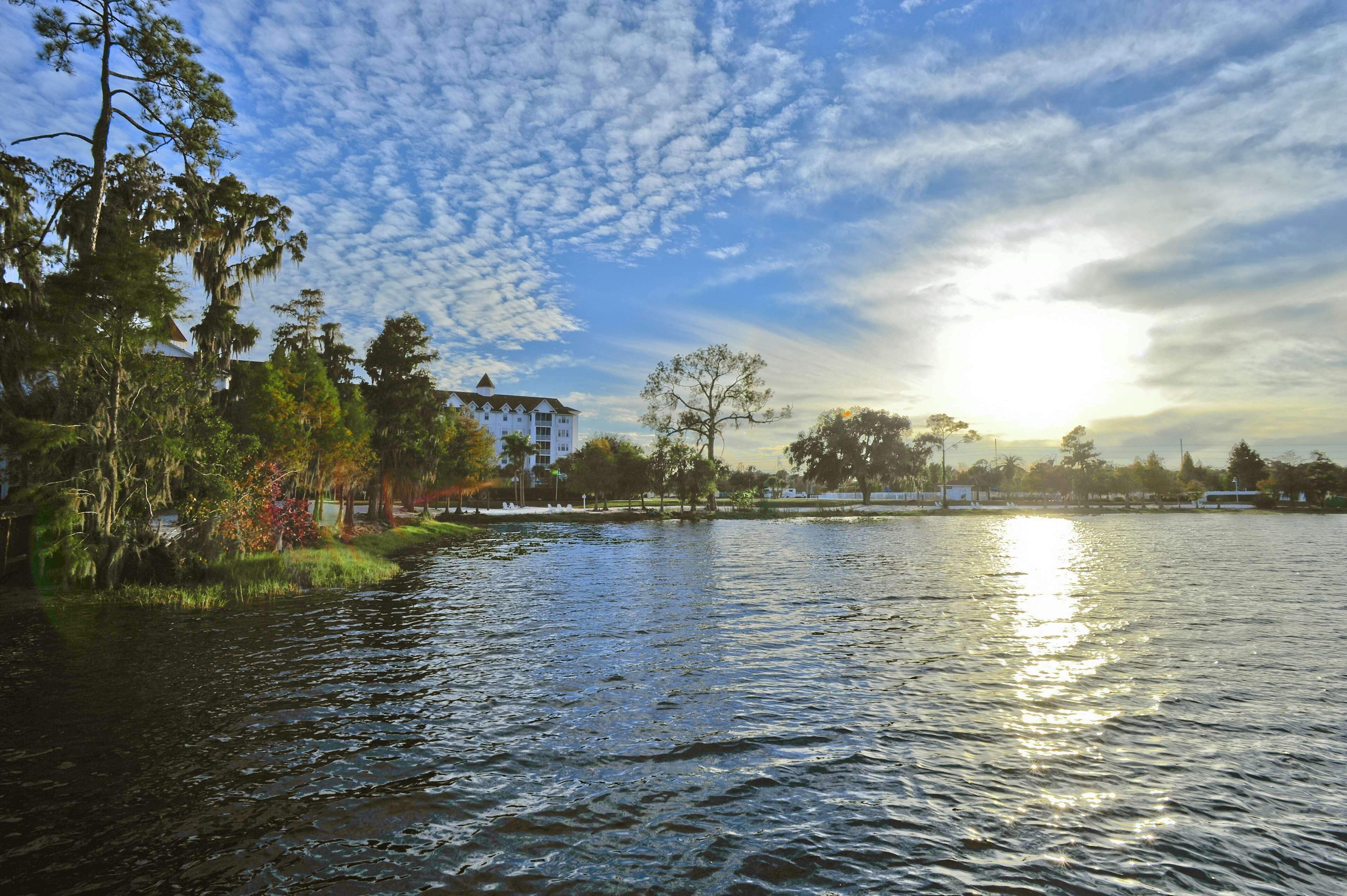 A calm river with a house on the left bank and trees in the background.