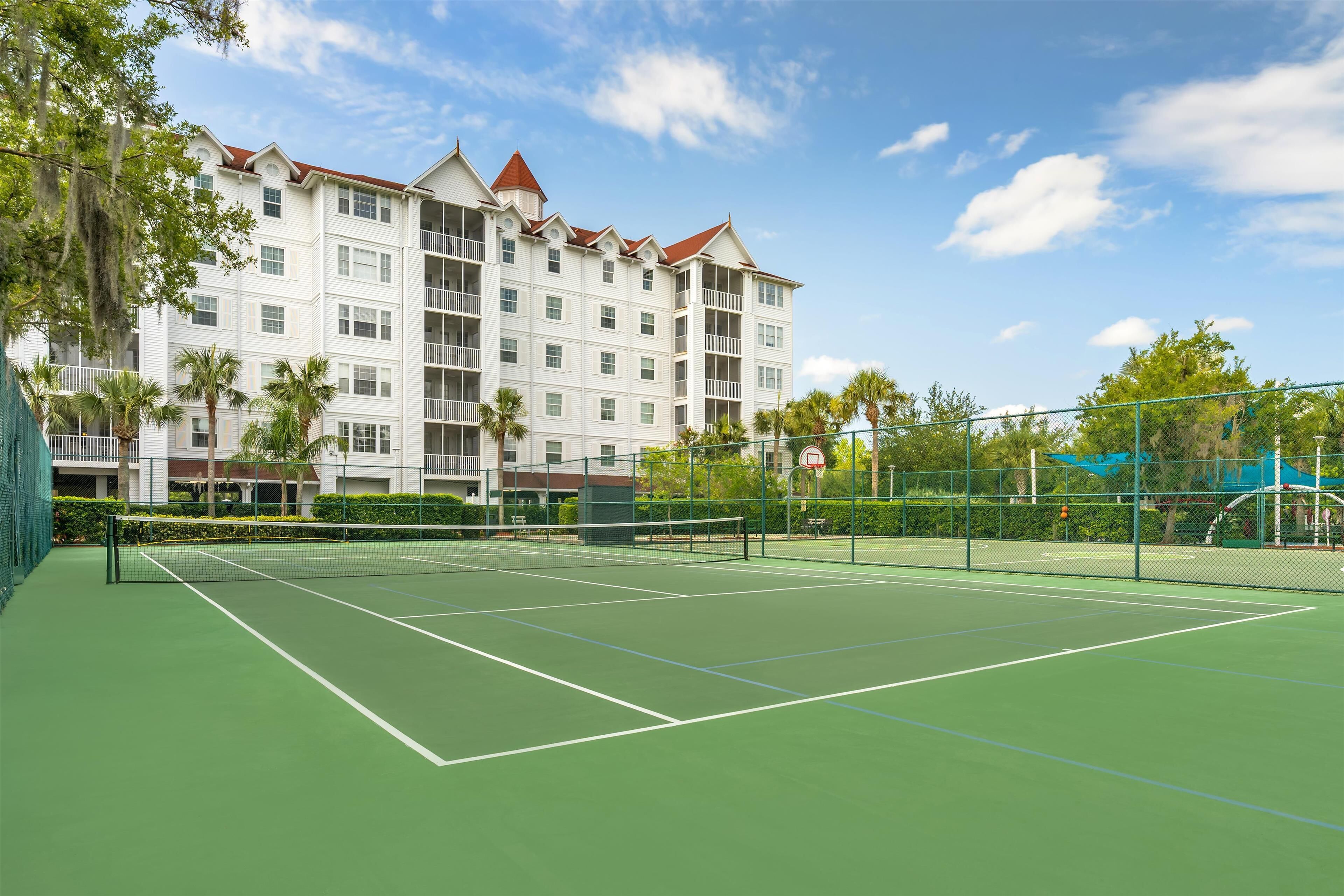 A tennis court with a net, surrounded by a fence, and a multi-story building with balconies in the background.