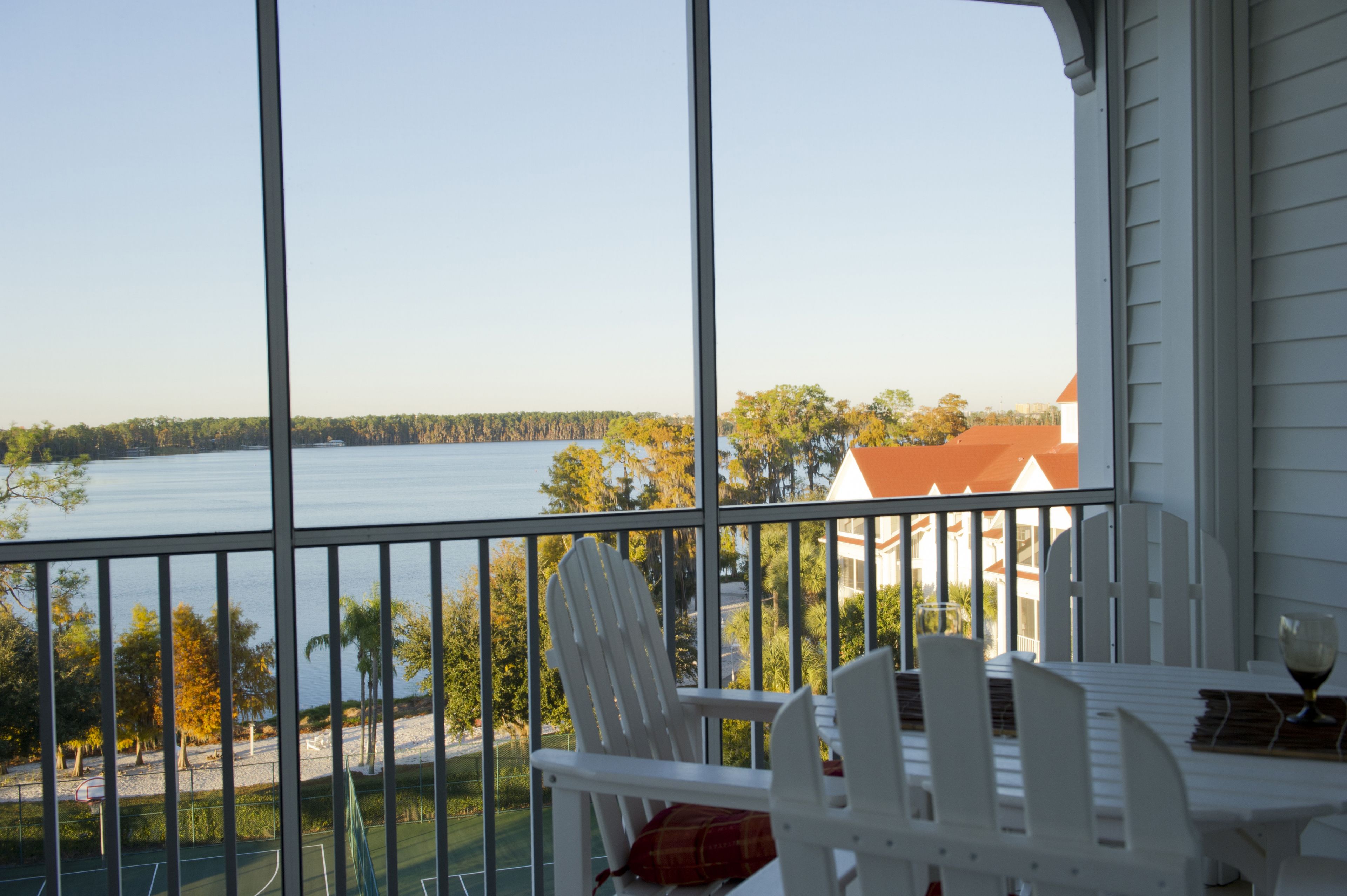 A balcony with a view of a lake, white chairs, and a table with a glass.