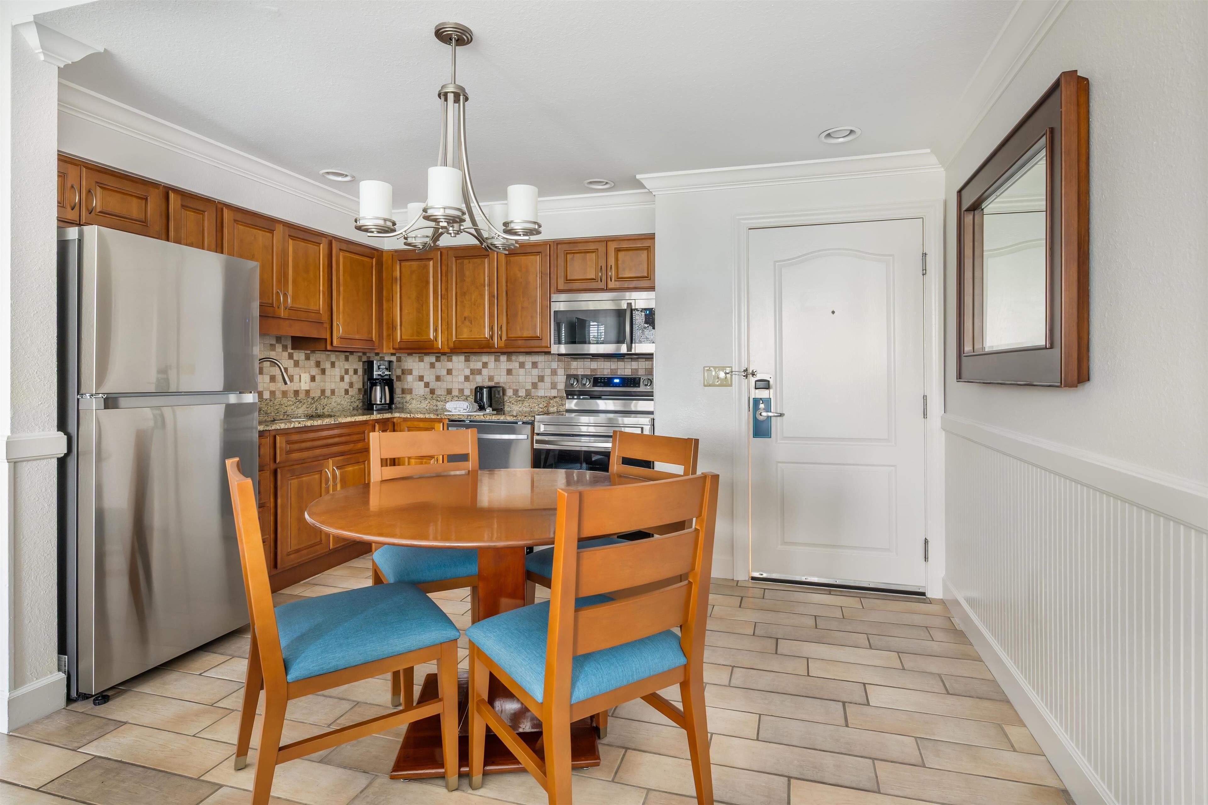A kitchen with wooden cabinets, a stainless steel refrigerator, a round dining table with blue upholstered chairs, and a hanging light fixture.