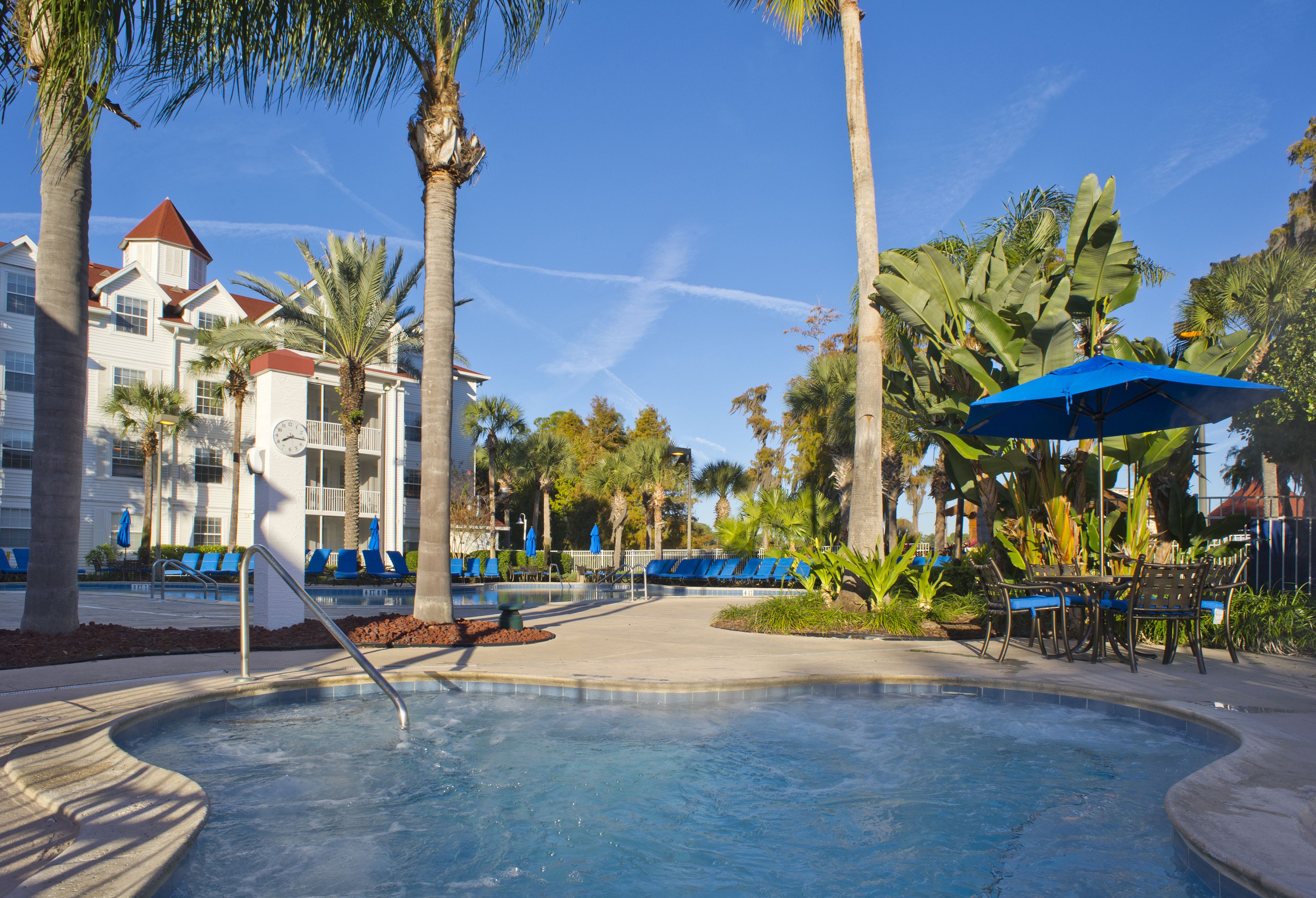 A pool area with lounge chairs and umbrellas, surrounded by palm trees and a building with a red roof.