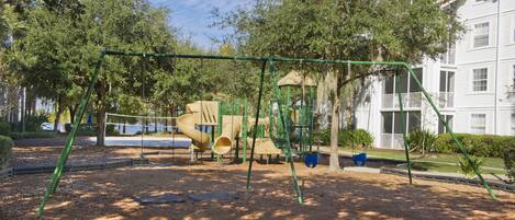 A playground with swings and slides, surrounded by trees and a building in the background.