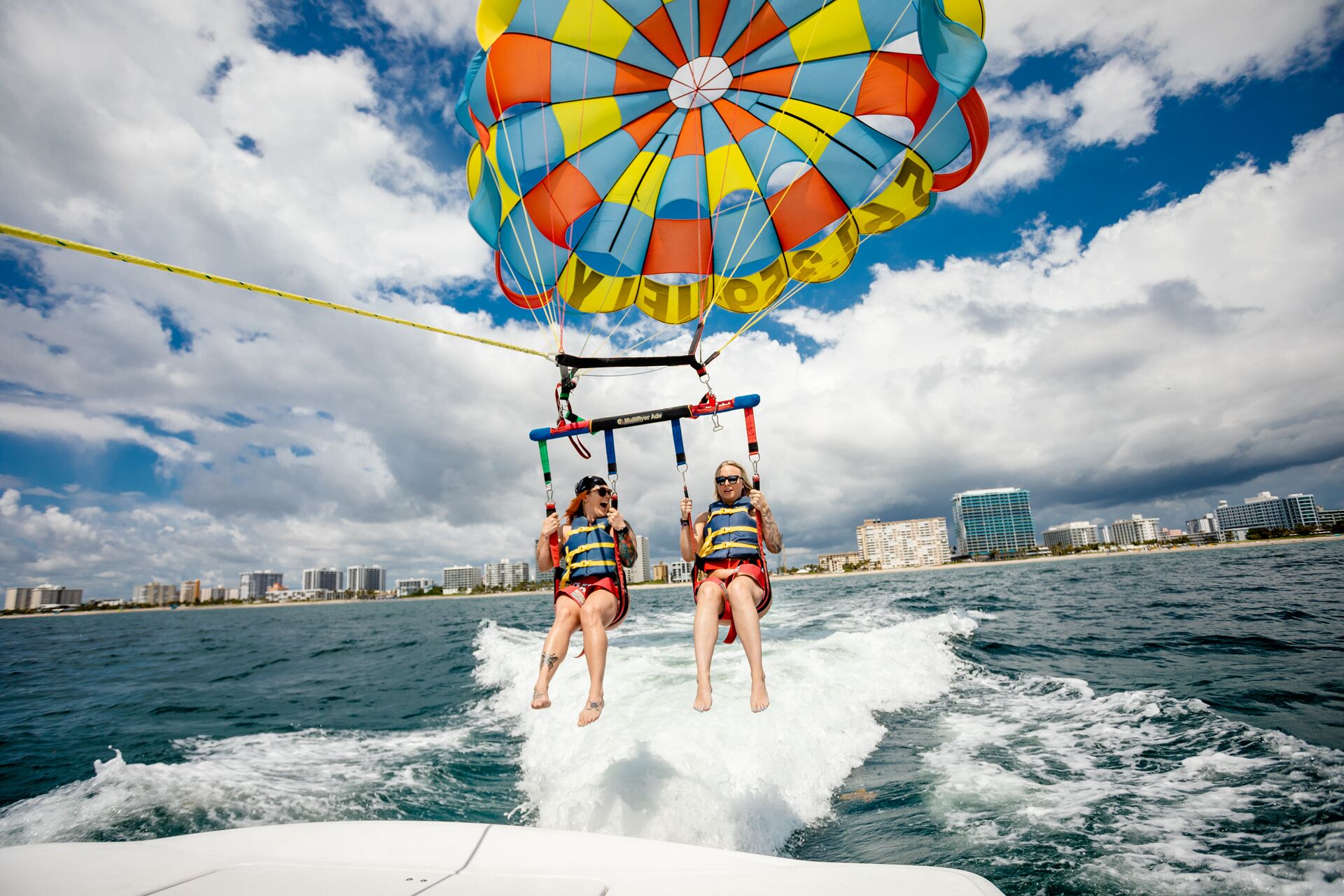 Two people parasailing over water with a city skyline in the background.