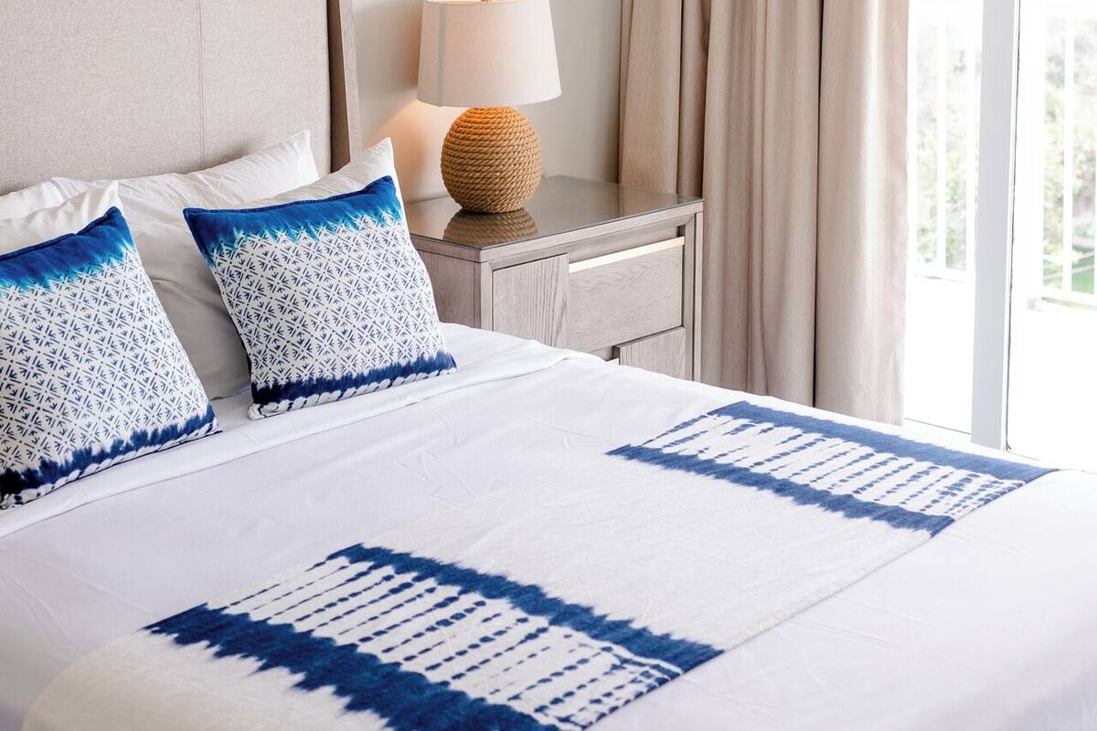 Habitación de hotel con una cama grande, una mesita de noche con lámpara y vistas al exterior.
