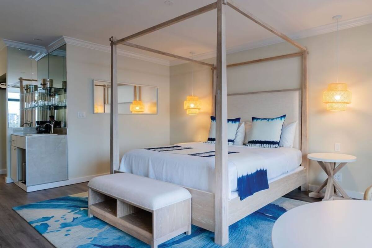 Una cama con dosel, un banco, mesitas de noche, una mesa redonda y una zona de cocina con un espejo.