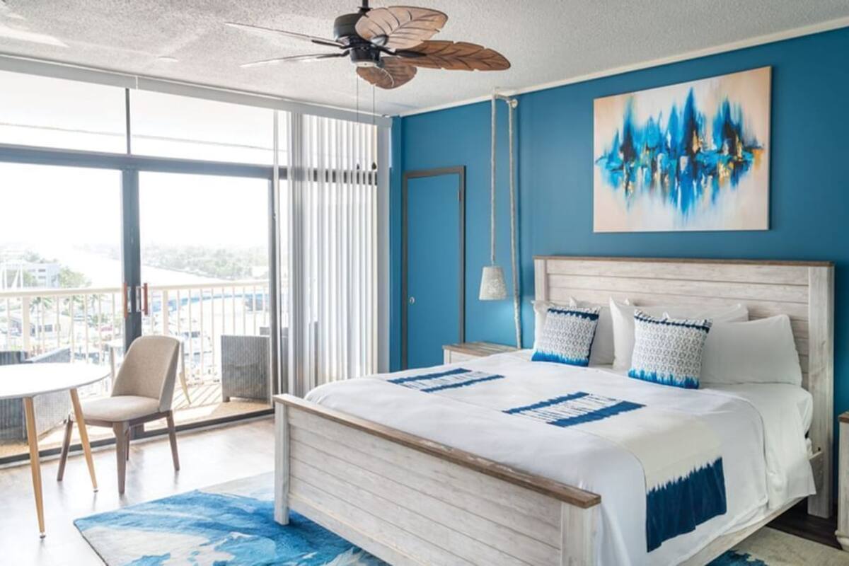 Un dormitorio con una cama grande, ventilador de techo, vistas de la ciudad y un balcón con mesa y silla.