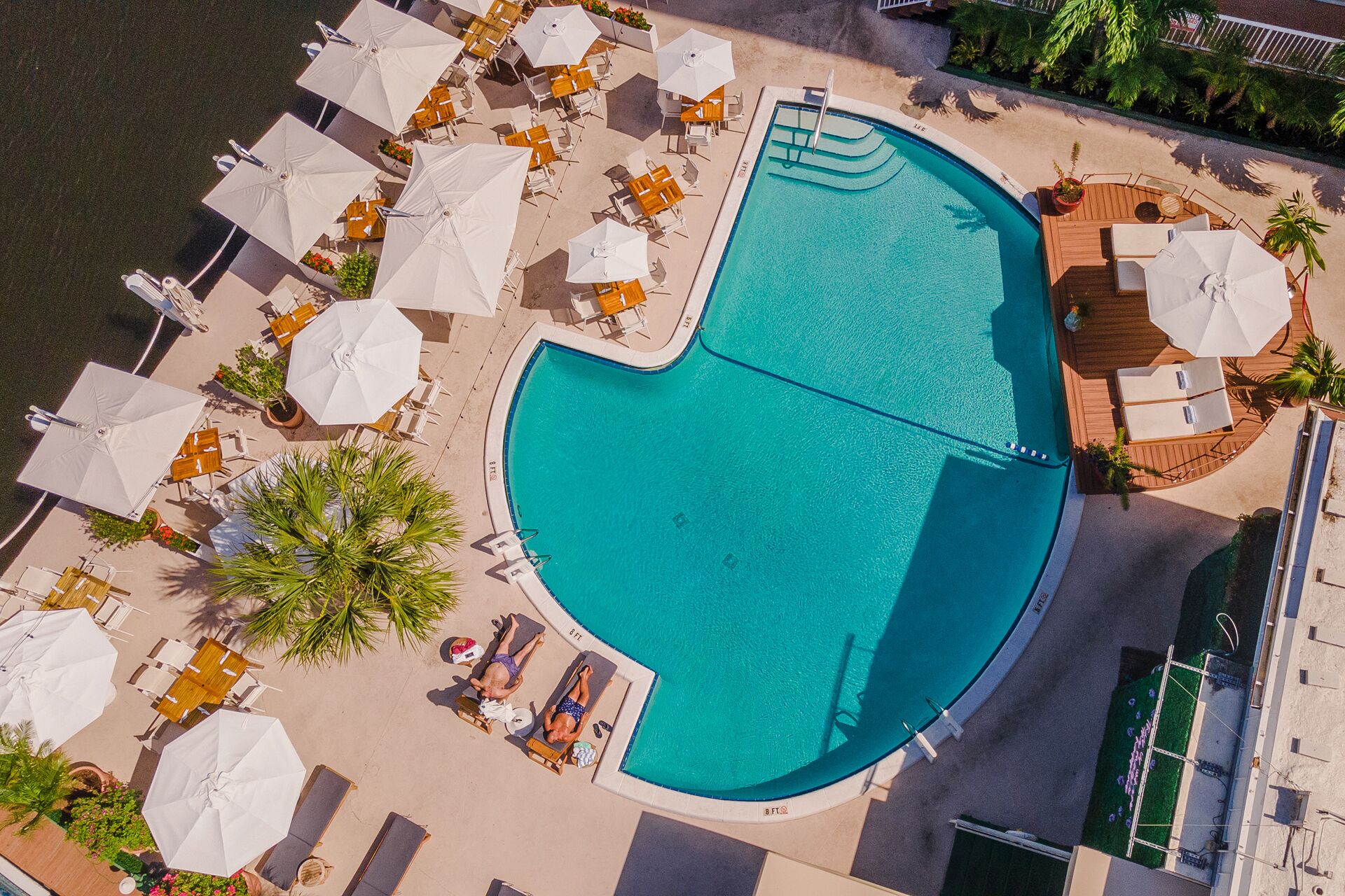 Vista aerea di una zona piscina con numerosi lettini e ombrelloni, circondata da un solarium sabbioso.