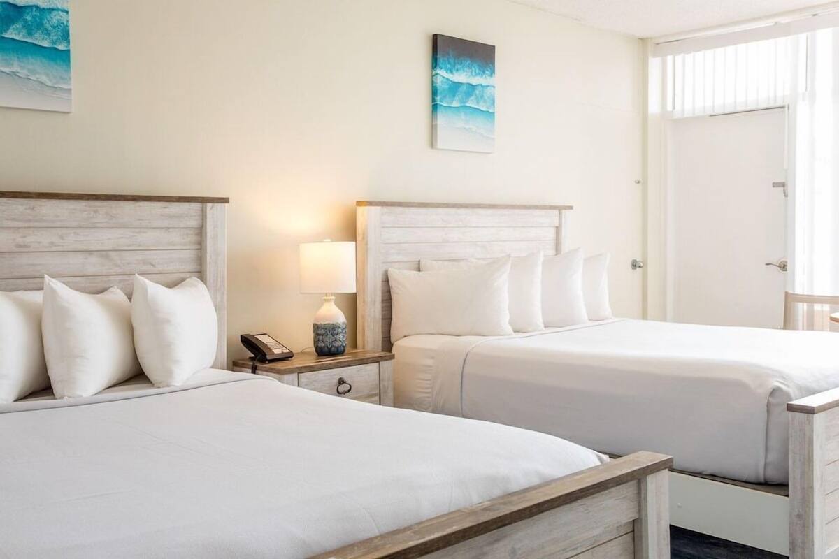 Habitación de hotel con dos camas, una mesita de noche con lámpara y un cuadro en la pared.