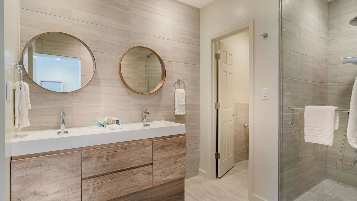 Une salle de bain moderne avec deux miroirs ronds, un meuble-lavabo en bois, une cabine de douche en verre et une porte donnant sur une autre pièce.