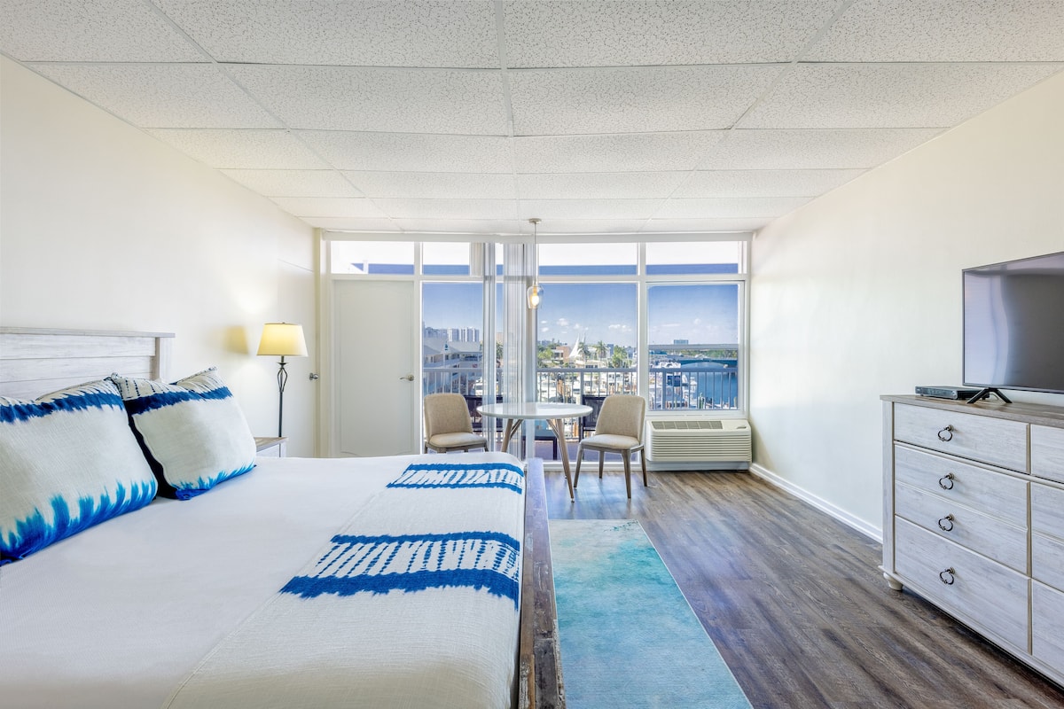 Habitación de hotel con una cama grande, televisión, escritorio con sillas y vistas a la ciudad a través de amplias ventanas.