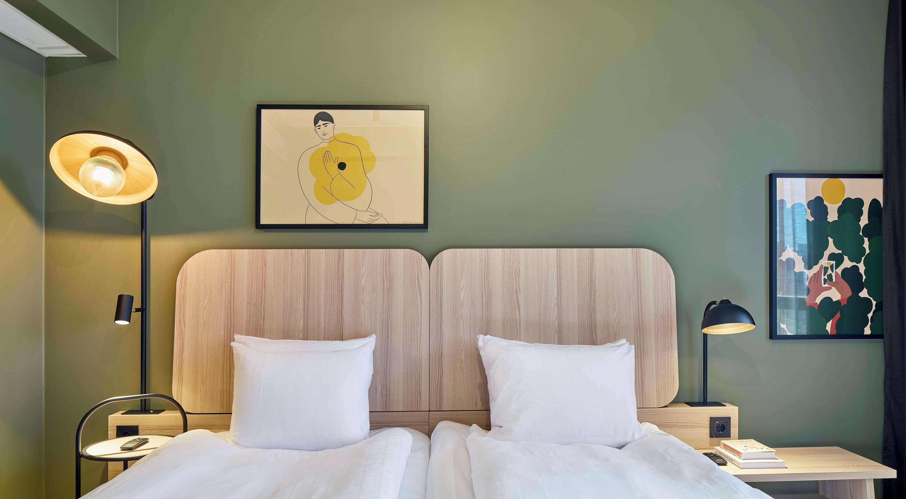 Habitación de hotel con cama doble, dos mesitas de noche, una lámpara de pared y dos cuadros enmarcados.