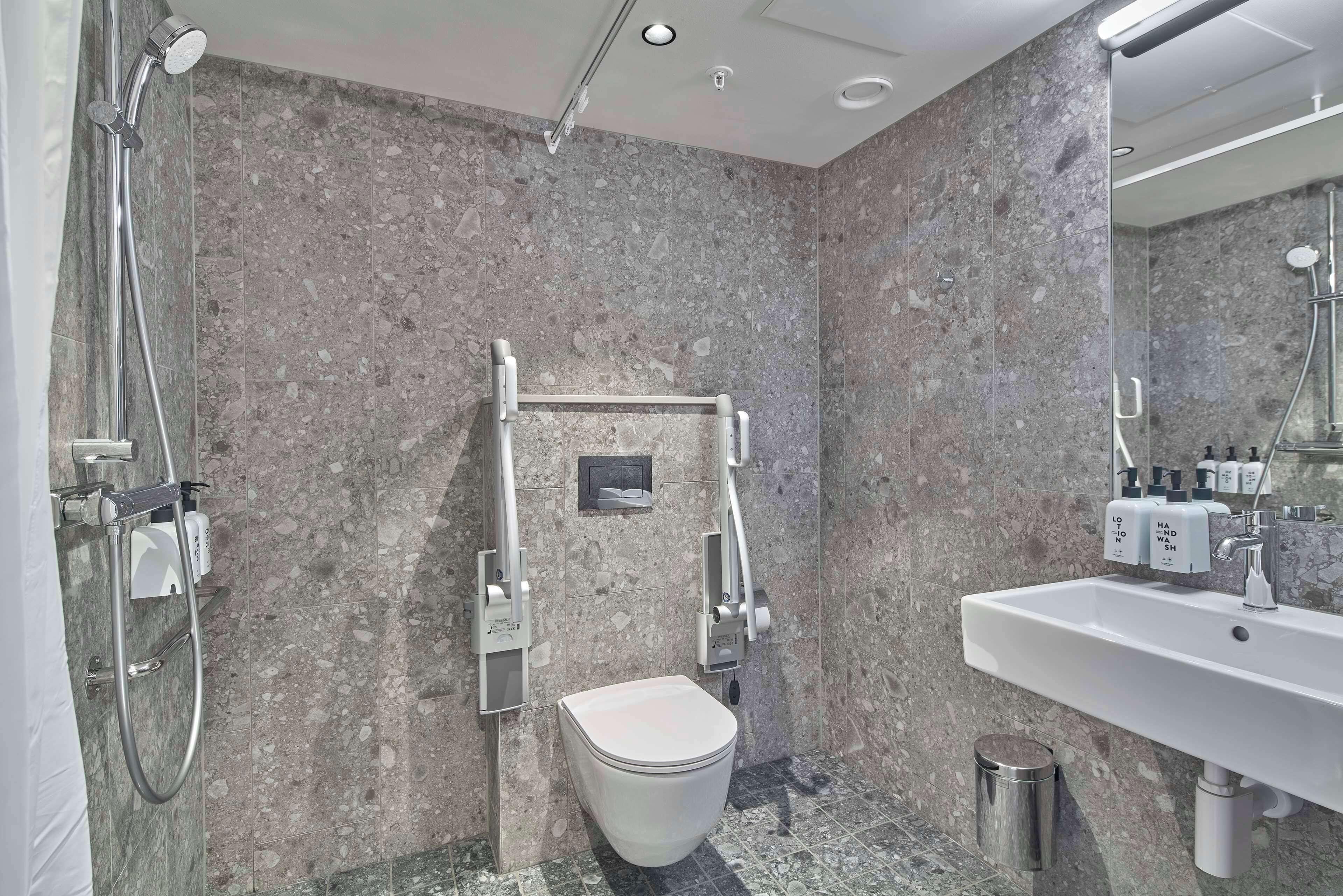 A modern bathroom with a marble floor, a white toilet, a shower with a handheld showerhead, a hand dryer, and a sink with a mirror.