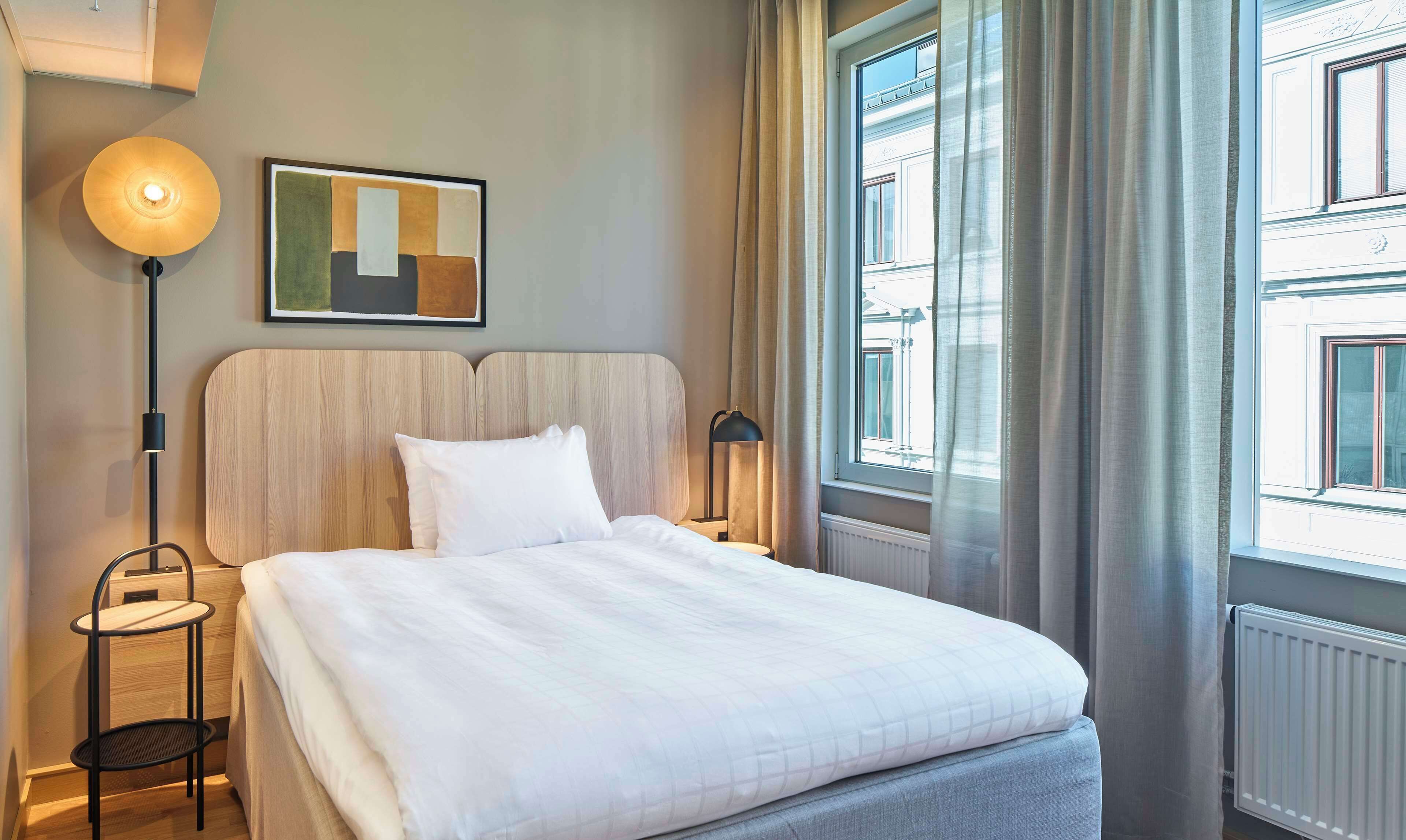 Une chambre d’hôtel avec un lit, une table de chevet, une lampe, un tableau et une fenêtre avec des rideaux.